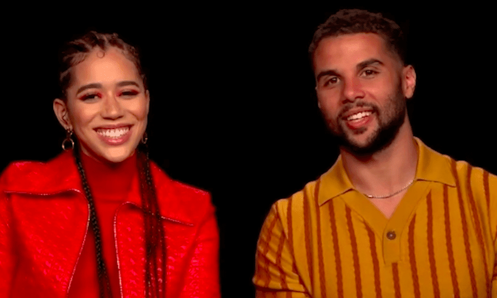 Two Scream VI cast members smile during an interview against a black background, wearing a bright red jacket and a mustard striped shirt.
