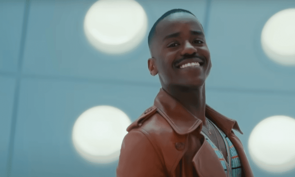 Ncuti Gatwa smiles as the Doctor in a brown jacket inside a bright futuristic room.