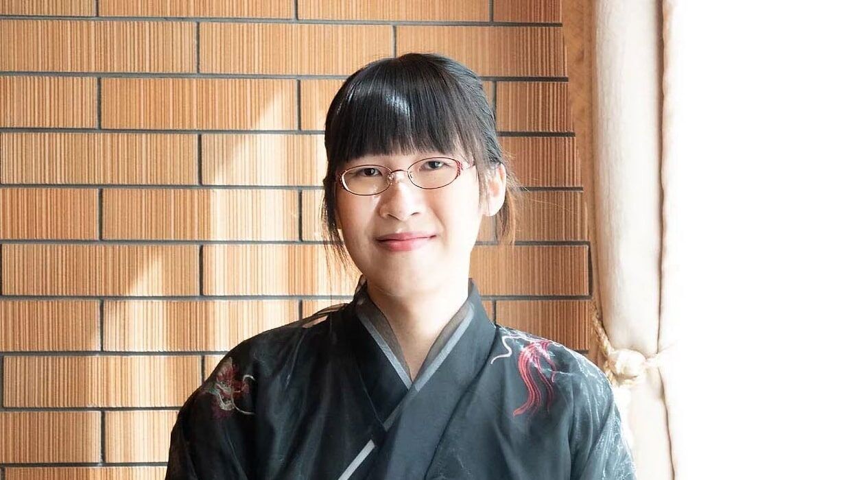 Novelist Li Kotomi wearing glasses and a dark kimono beside a brick wall.