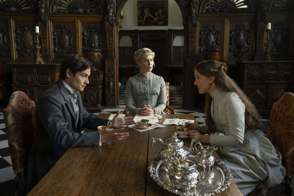 "Trois personnes vêtues de vêtements de l'époque victorienne sont assises autour d'une table en bois avec un service à thé en argent dans une pièce ornée de lambris et discutent."