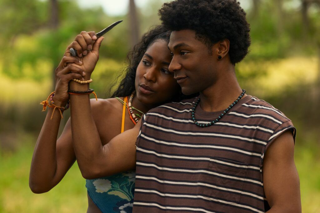 "Two people stand closely together outdoors. The woman holds up a knife while the man gently guides her hand. They both appear calm and focused, surrounded by lush greenery in a natural setting."