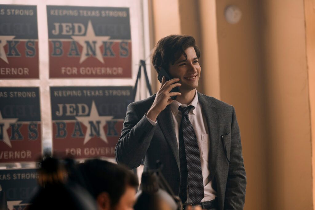 "Jeune homme en costume souriant tout en parlant au téléphone dans un bureau décoré d'affiches de campagne politique qui disent "Jed Banks pour le gouverneur" et "Garder le Texas fort." Éclairage intérieur chaleureux avec d’autres personnes travaillant en arrière-plan."