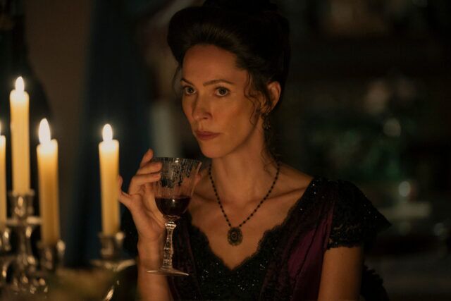 "Une femme en costume d'époque est assise à une table éclairée aux chandelles, tenant un verre de vin rouge, avec une expression sérieuse dans une pièce élégante et faiblement éclairée."