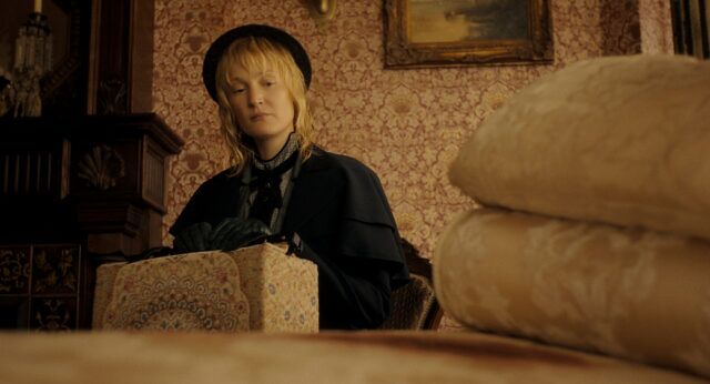 "A woman in vintage clothing and gloves sits indoors, holding a patterned box. The room has ornate wallpaper, a painting, and folded linens in the foreground, suggesting a historical or period setting."