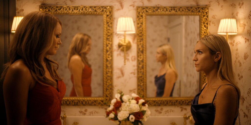 "Deux femmes vêtues de robes élégantes se font face dans une salle de bains chaleureusement éclairée et ornée de papier peint à fleurs, de miroirs aux cadres dorés, d'appliques murales et d'un vase de fleurs sur le comptoir, leurs reflets étant visibles dans les miroirs."